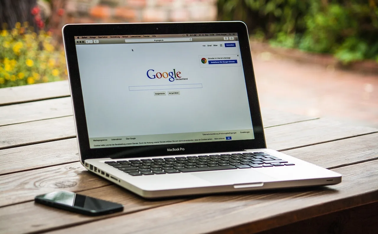 Laptop displaying Google search page on a wooden table, with a smartphone nearby, reflecting a modern workspace.