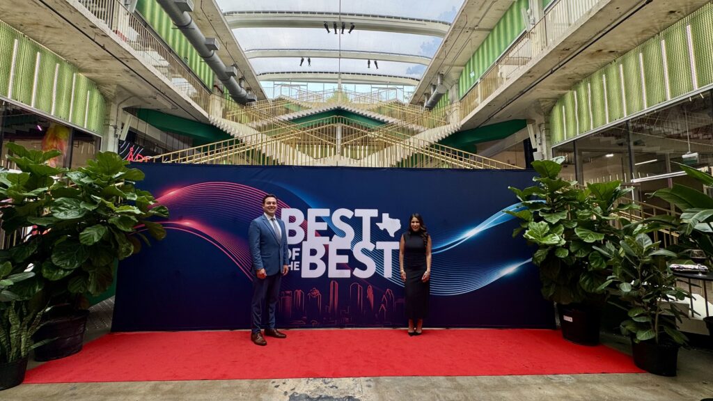 David Smith Law Firm representatives posing in front of a "Best of the Best" backdrop at a celebratory event, surrounded by greenery in a modern venue.